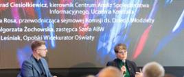Na zdjęciu siedzą od lewej: Konrad Ciesiołkiewicz i płk Małgorzata Żochowska. W tle ścianka multimedialna. Na pierwszym planie na dole zdjęcia głowy innych uczestników konferencji.
