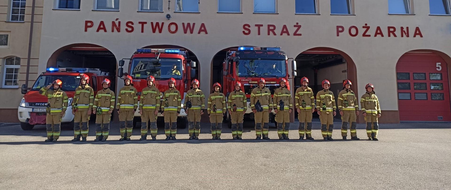 Strażacy ustawieni w szeregu przed pojazdami z załączoną sygnalizacją