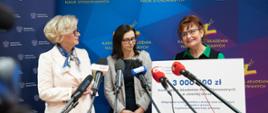 Wiceminister Machałek w białym ubraniu stoi obok kobiety w szarym sweterku i ubranej na zielono kobiety trzymającej wielki symboliczny czek z napisem 3 000 000 zł, przed nimi kilka mikrofonów na stojakach, za nimi niebieska ścianka z napisem Karkonoska Akademia Nauk Stosowanych.