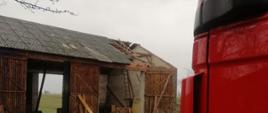 Na zdjęciu widać budynek stodoły. Wrota otwarte. Uszkodzony dach w szczycie. Po prawej fragment samochodu ratowniczo-gaśniczego. Kolor samochodu czerwony. Zachmurzenie duże.