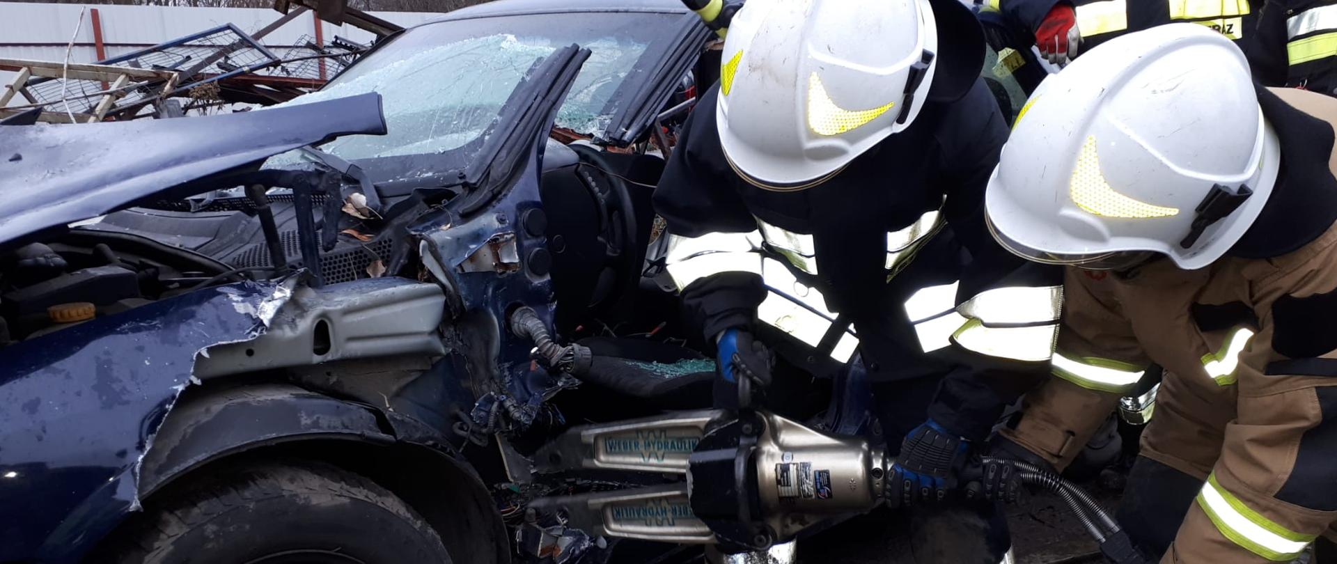 Dwaj strażacy ubrani w ubrania specjalne i hełmy, pochyleni, trzymają nożyce hydrauliczne którymi rozpierają karoserię samochodu w okolicy zawiasów drzwi przednich samochodu osobowego.