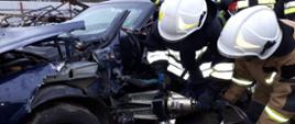 Dwaj strażacy ubrani w ubrania specjalne i hełmy, pochyleni, trzymają nożyce hydrauliczne którymi rozpierają karoserię samochodu w okolicy zawiasów drzwi przednich samochodu osobowego.