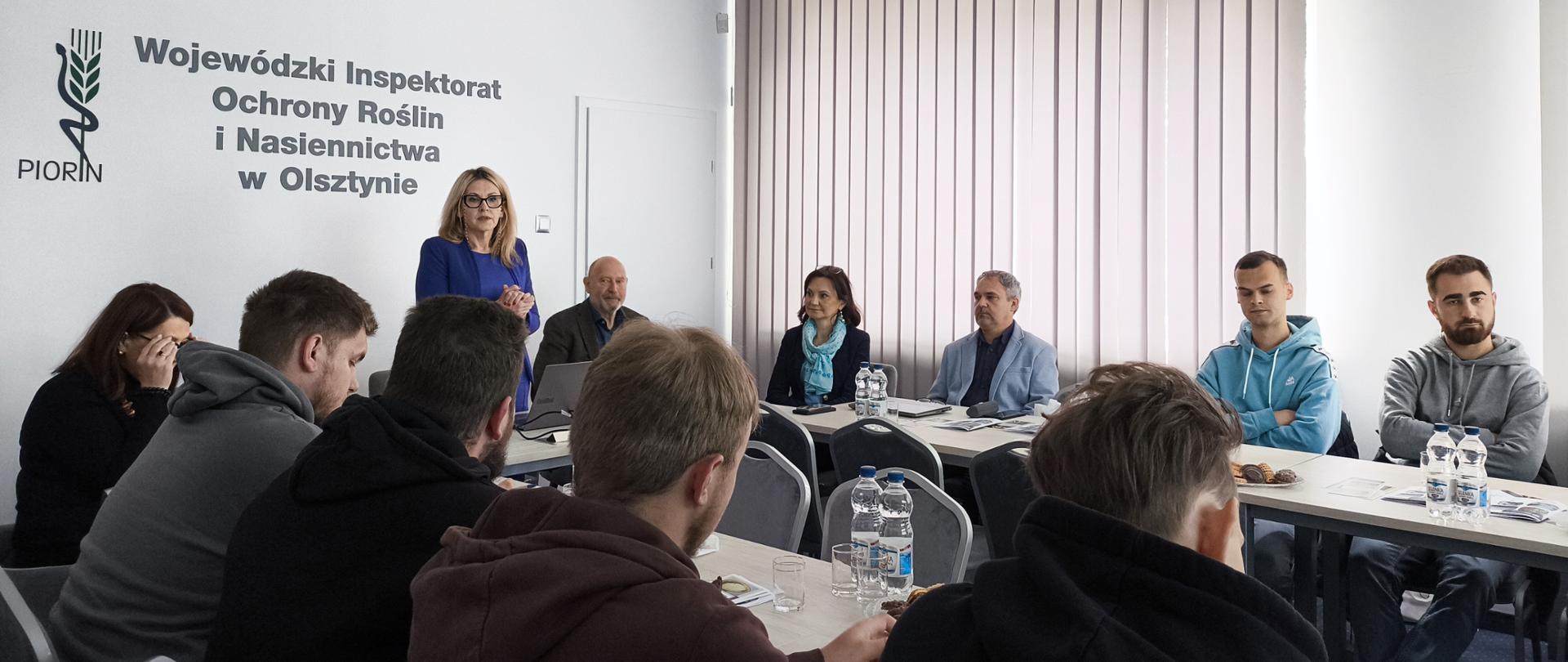 w głębi zdjęcia stoi stoi kobieta i mówi do osób siedzących przy stołach ustawionych naprzeciw siebie