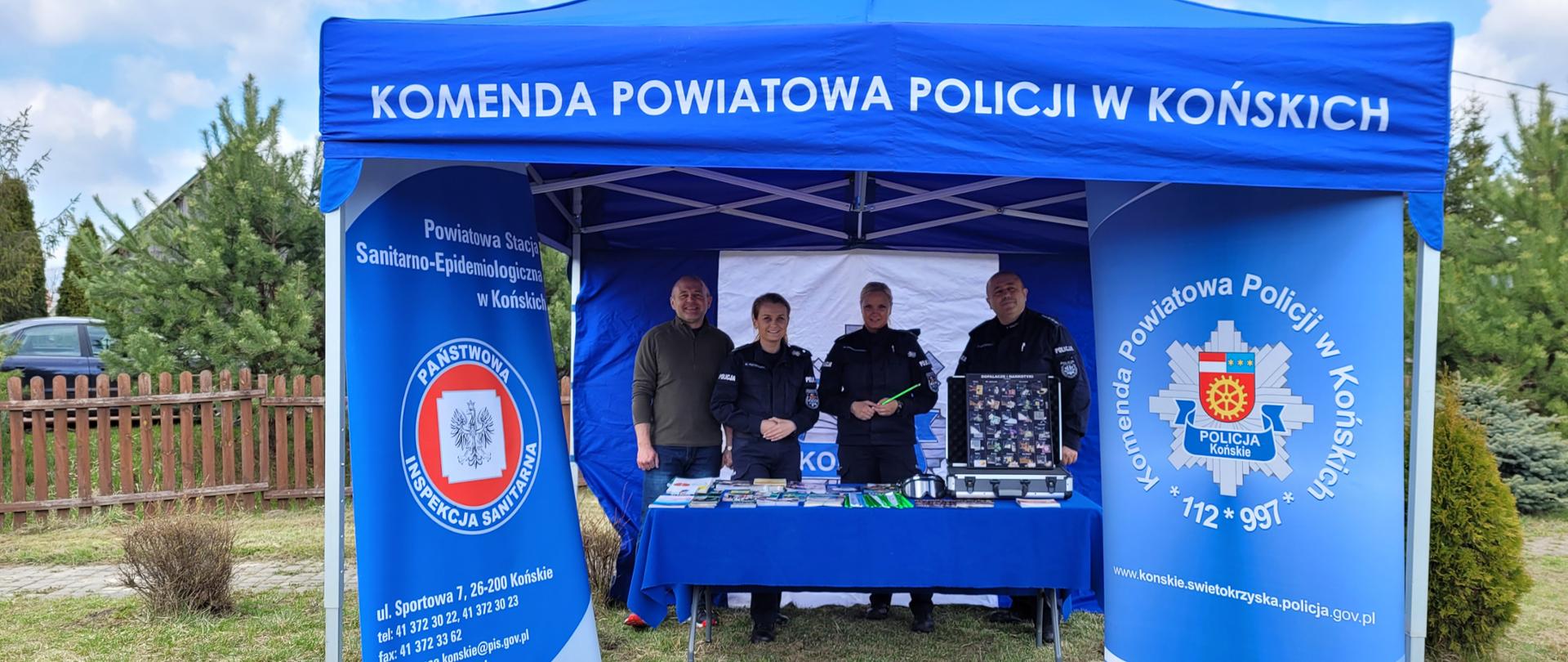 Niebieski namiot wystawowy z napisem u góry: KOMENDA POWIATOWA POLICXJI W KOŃSKICH. Po bokach namiotu 2 rollapy reklamowe z godłami: Policji i Inspekcji Sanitarnej. Pod namiotem troje policjantów w mundurach - dwie kobiety i mężczyzna oraz w stroju cywilnym pracownik Inespekcji Sanitarnej w Końskich. W tle tereny zielone.