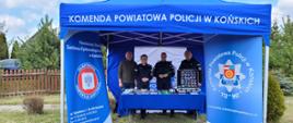 Niebieski namiot wystawowy z napisem u góry: KOMENDA POWIATOWA POLICXJI W KOŃSKICH. Po bokach namiotu 2 rollapy reklamowe z godłami: Policji i Inspekcji Sanitarnej. Pod namiotem troje policjantów w mundurach - dwie kobiety i mężczyzna oraz w stroju cywilnym pracownik Inespekcji Sanitarnej w Końskich. W tle tereny zielone.