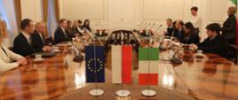 Przy wielkim owalnym drewnianym błyszczącym stole siedzą uczestnicy spotkania, przed każdym mikrofon, z przodu na stole stoją małe flagi UE, Polski i Włoch, z lewej strony za stołem dwa okna.