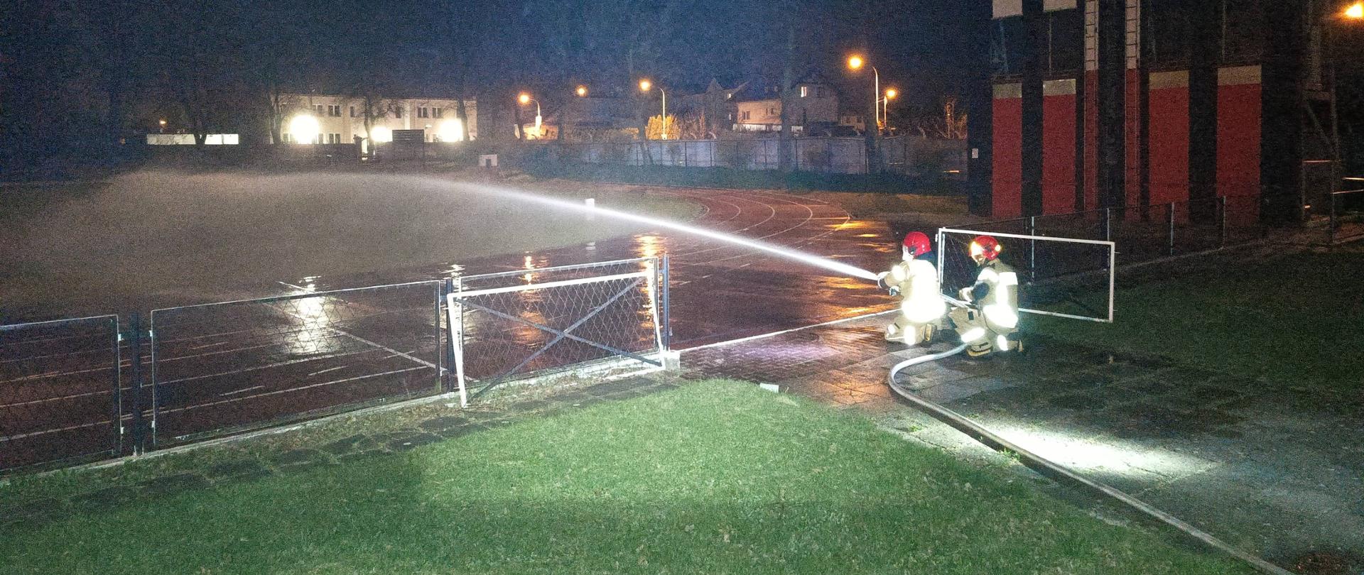 Nocne ćwiczenia, rota strażaków kieruje strumień wody na teren boiska, w głębi po prawej widoczna wspinalnia