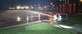 Nocne ćwiczenia, rota strażaków kieruje strumień wody na teren boiska, w głębi po prawej widoczna wspinalnia