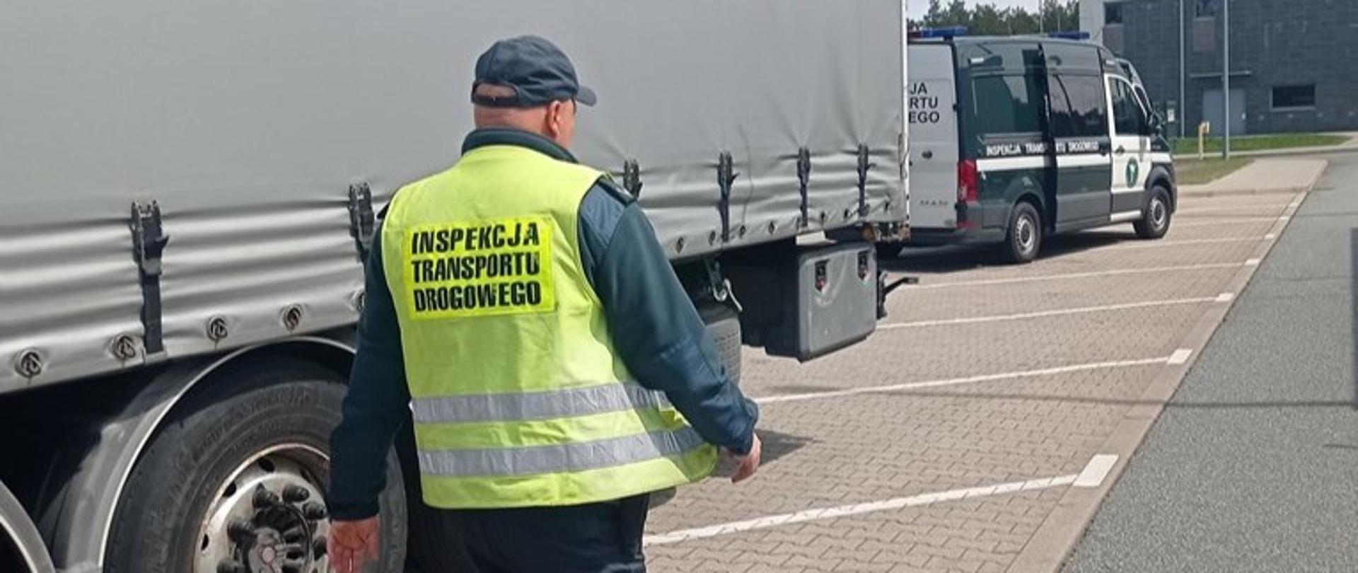 Na pierwszym planie: umundurowany inspektor lubuskiej Inspekcji Transportu Drogowego przechodzi obok naczepy kontrolowanego zestawu ciężarowego. Za pojazdem ciężarowym stoi oznakowany furgon Inspekcji Transportu Drogowego.