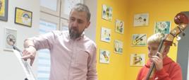 Na zdjęciu znajdują się pedagog prowadzący warsztaty - Wojciech Front oraz uczeń z kontrabasem w sali rytmicznej PSM w kolorach szaro, żółty, biały, na ścianach wiszą kolorowe obrazki muzyczne