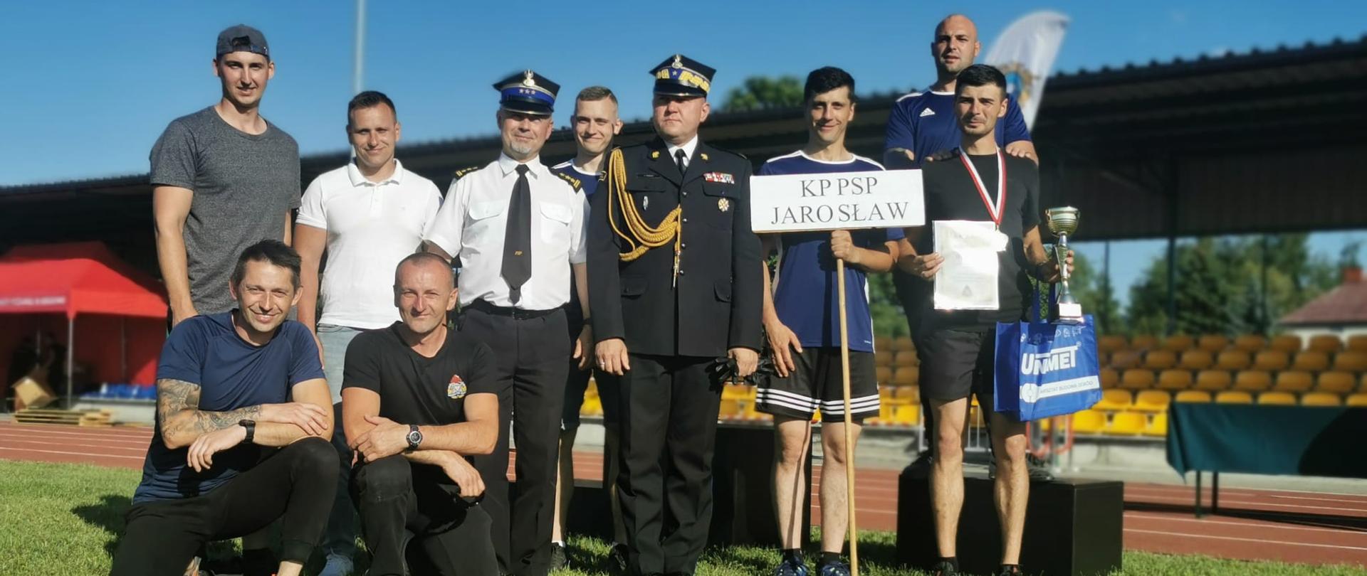 Na zdjęciu widzimy reprezentację KP PSP Jarosław wraz z Podkarpackim Komendantem Wojewódzkim PSP oraz Komendantem Powiatowym PSP w Jarosławiu. Zawodnicy ubrani są w strój sportowy, a komendanci w umundurowanie wyjściowe. Jeden ze strażaków trzyma puchar i torbę z nagrodami, a drugi tablicę z nazwą komendy.