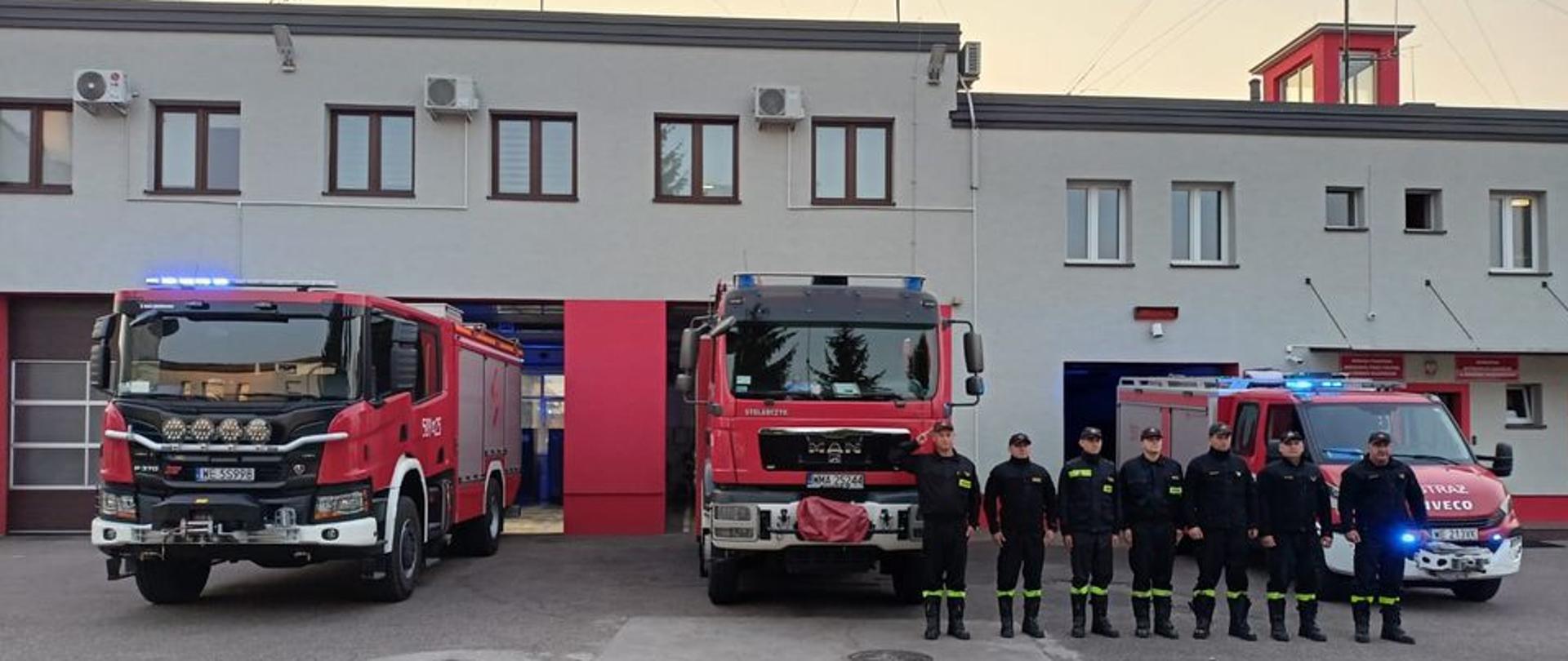 Strażacy stoją w szeregu przed remizą. W samochodach widać włączone sygnały świetlne. 