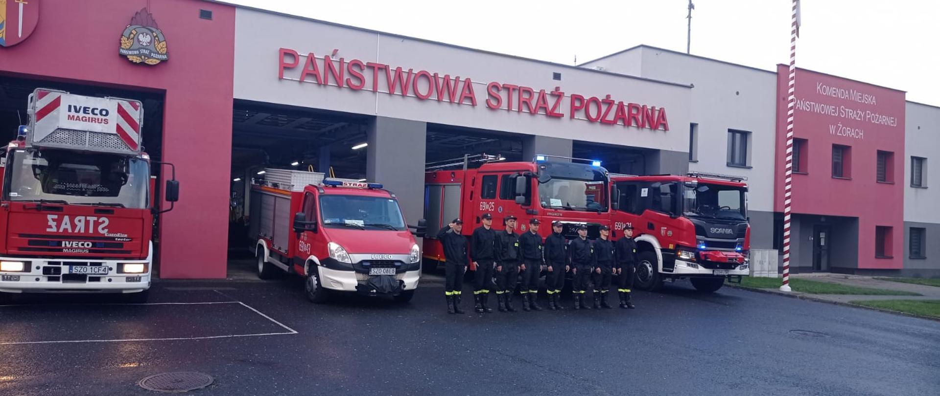 Strażacy w szeregu stoją na tle samochodów pożarniczych wystawionych do połowy z garażu. W prawej części zdjęcia widoczny budynek Komendy. Nad głowami strażaków napis PAŃSTWOWA STRAŻ POŻARNA oraz logo PSP.
