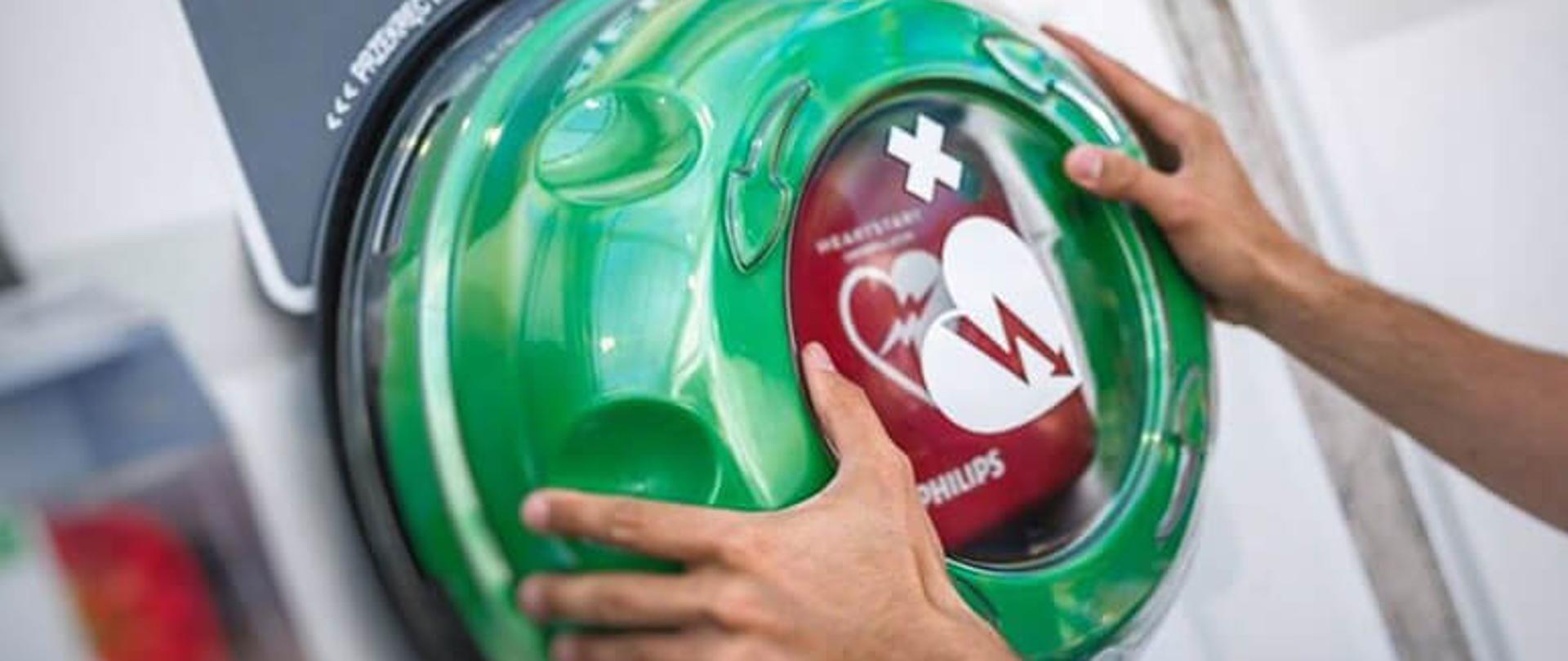 Dłonie chwytające zielone opakowanie z napisem AED, powyżej tablica z napisem defibrylator. 