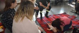 Szkolenie z pierwszej pomocy. Świetlica Komendy Powiatowej Policji w Rawiczu. Na pierwszym planie jedna z uczestniczek szkolenia, która klęcząc - pochyla się nad leżącym na kocu manekinie "niemowlaku" - wykonując czynności ratujące życie (masaż serca i sztuczny oddech) po uprzednim nagłym zatrzymaniu krążenia u dziecka. Wokół stanowiska ćwiczebnego znajdują się inne uczestniczki zajęć i strażak-instruktor.