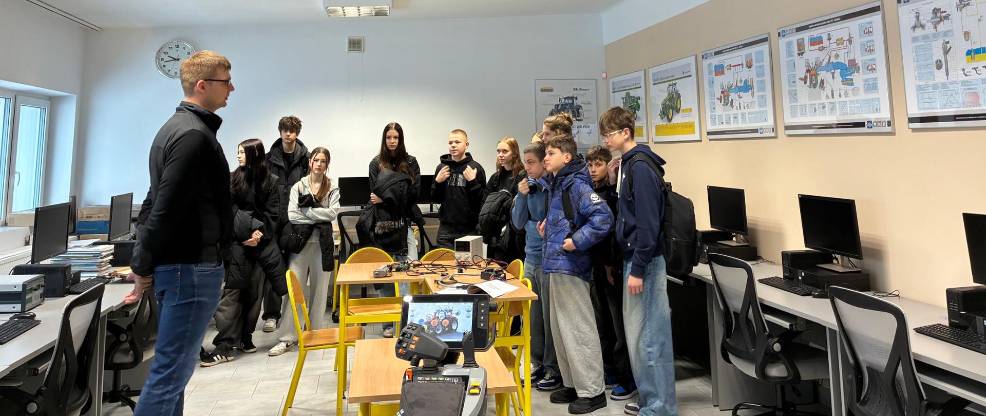Młodzież w pracowni komputerowej słucha wykładu nauczyciela stojącego po lewej stronie, z widocznym na stole elementem sprzętu symulatora lub robota i plakatami technicznymi na ścianie.