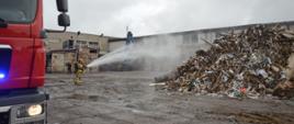 Akcja na składowisku odpadów. Plac, na którym znajdują się pryzmy z odpadami. Na pierwszym planie fragment wozu straży pożarnej oraz strażak gaszący pożar odpadów przy użyciu wody. Na ziemi znajduje się rozwinięty wąż strażacki koloru białego. w tle zabudowania firmowe.