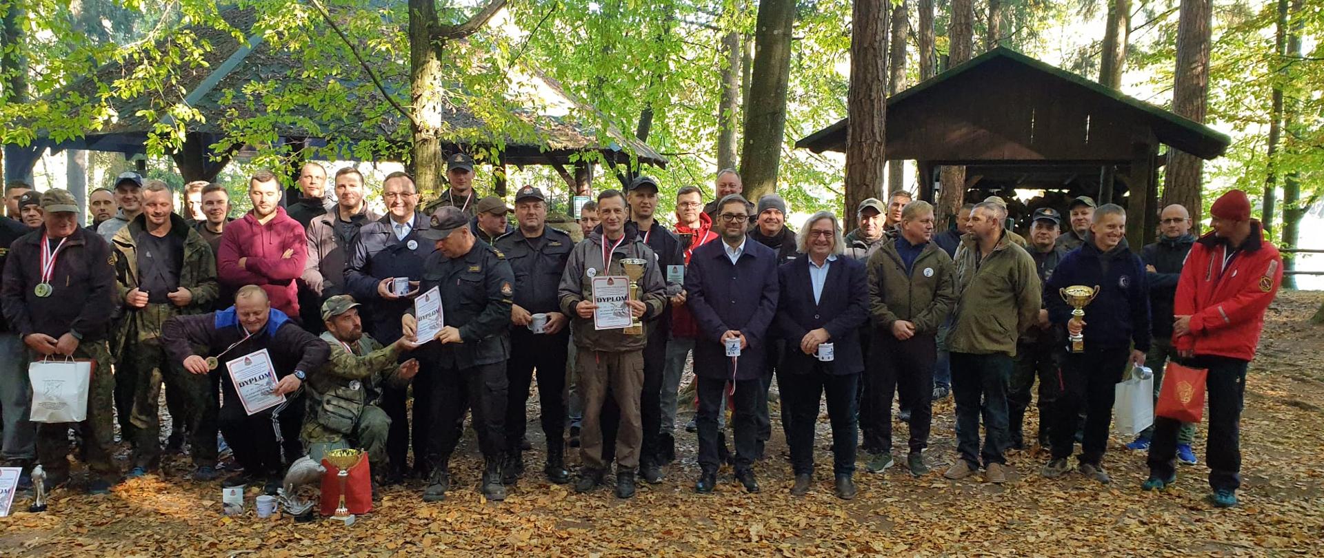 Na zdjęciu wszyscy uczestnicy zawodów wędkarskich. W tle wiaty drewniane. Piękna słoneczna jesienna pogoda.