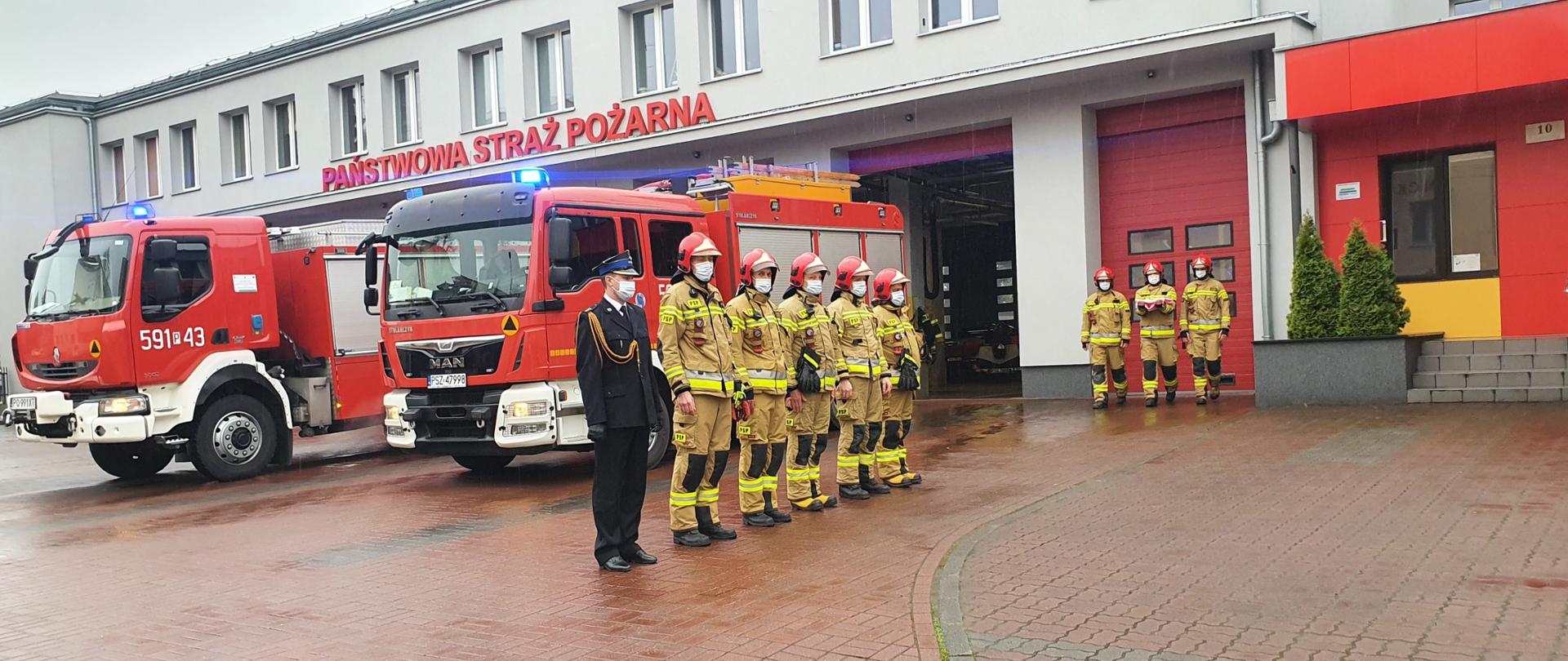 Trzech strażaków niesie flagę w celu wciągnięcia jej na maszt. Przed budynkiem Straży Pożarnej stoją samochody oraz inni strażacy.