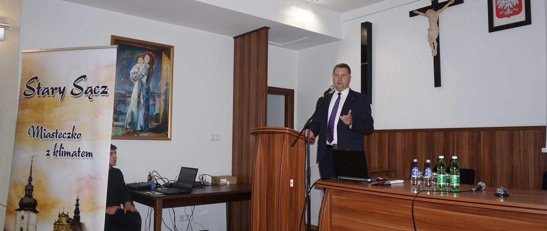 Zdjęcie z boku, minister Czarnek stoi za drewnianą mównicą i mówi do mikrofonu, z przodu baner z napisem Stary Sącz - miasteczko z klimatem.