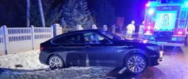 Na zdjęciu wykonanym w porze nocnej widać uszkodzony pojazd. Oświetlenie sztuczne. Zdjęcie na drodze publicznej. Pozostałości śniegu na poboczu. W kadrze znajduje się również samochód ratowniczo-gaśniczy w kolorze czerwonym z sygnalizacją ostrzegawczą.
