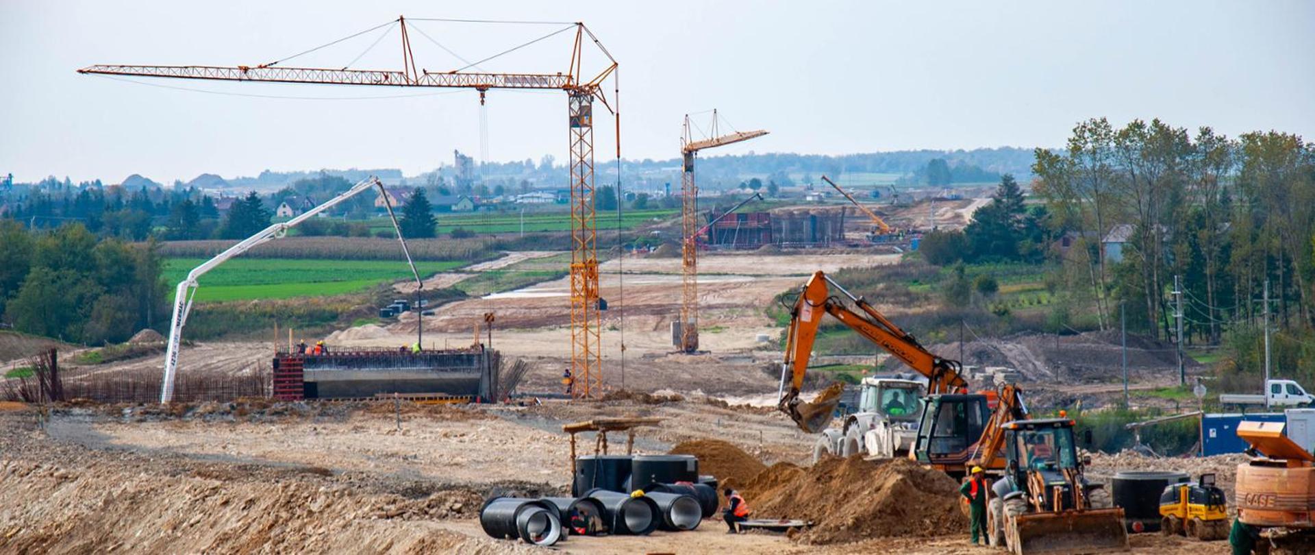 Na zdjęciu przedstawiono plac budowy drogi ekspresowej w otwartym terenie. W centralnej części kadru widoczne są duże maszyny budowlane, w tym koparka wykonująca prace ziemne oraz walec drogowy przygotowujący podłoże. Obok nich znajdują się pracownicy w odzieży roboczej, którzy nadzorują prace.
W tle rozciąga się szeroki pas przygotowywanego nasypu, na którym widać kilka wysokich żurawi wieżowych oraz betonowe konstrukcje – prawdopodobnie elementy przyszłych wiaduktów lub mostów. Na dalszym planie widoczne są zielone pola i drzewa, co podkreśla kontrast między naturalnym krajobrazem a intensywnymi pracami budowlanymi.
Kolorystyka zdjęcia jest naturalna, z dominacją odcieni ziemi (brązy, beże) oraz zieleni w tle. Niebo jest jasne, lekko zachmurzone, co sugeruje neutralne warunki pogodowe sprzyjające pracom.