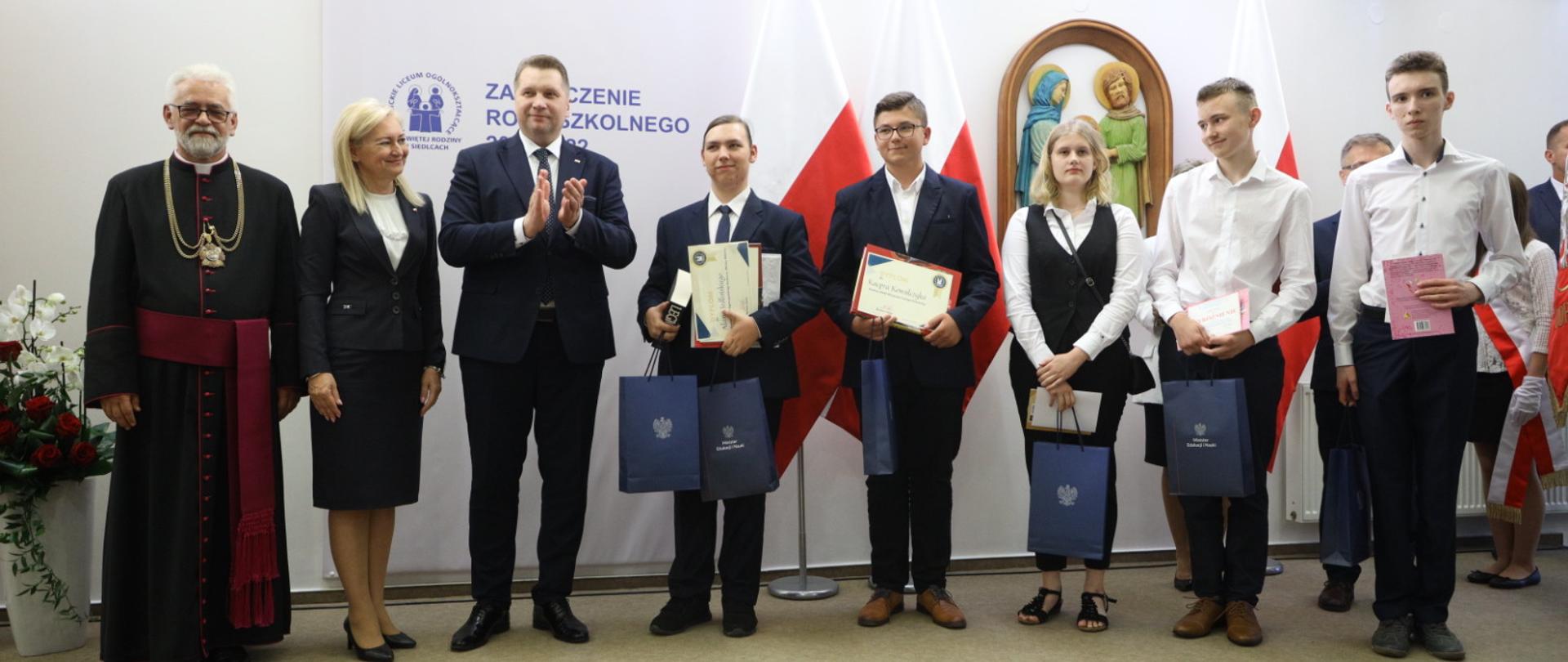 Zdjęcie zbiorowe uczestników uroczystości, personel szkoły oraz minister Czarnek stoją pod ścianą, kilkoro uczniów pokazuje dyplomy.