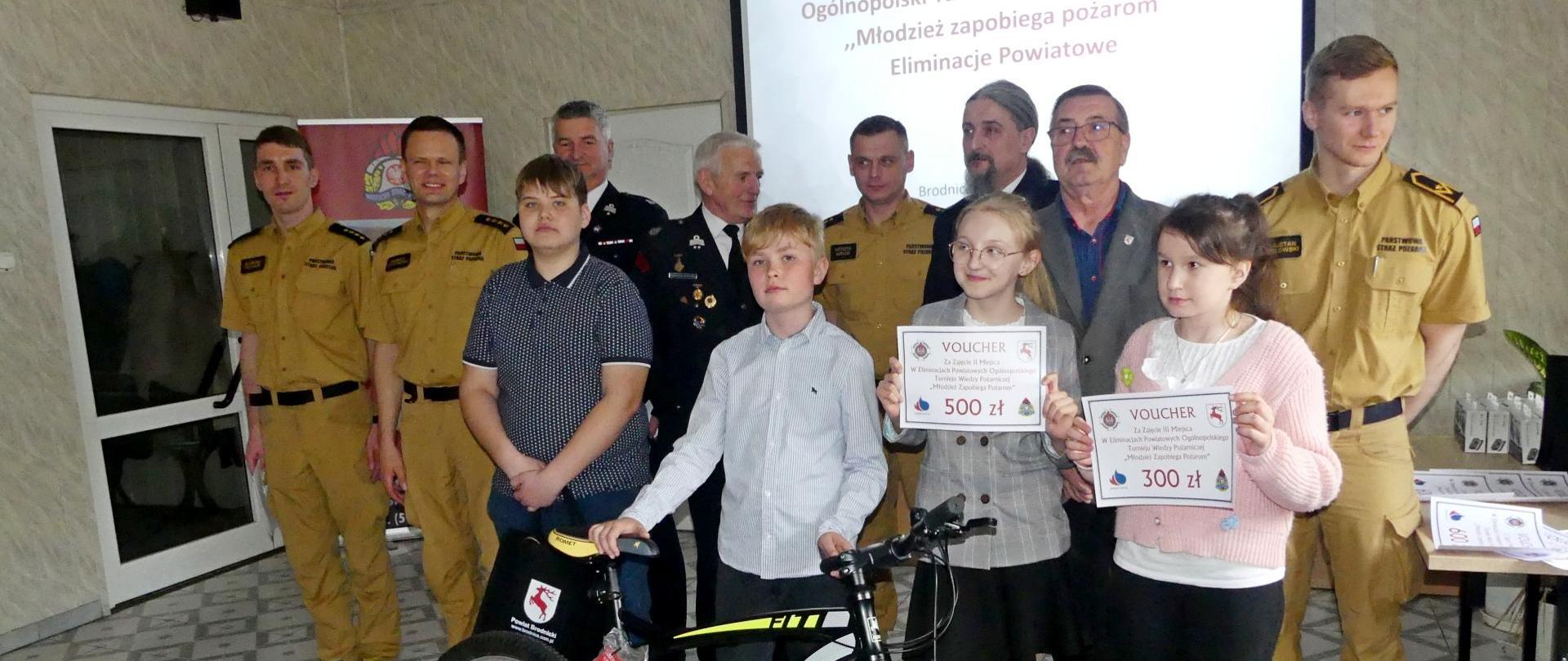 Zdjęcie przedstawia zwycięzców w najmłodszej grupie wiekowej eliminacji szczebla powiatowego Ogólnopolskiego Turnieju Wiedzy Pożarniczej „Młodzież zapobiega pożarom”. Za zwycięzcami przedstawiciele organizatorów. Zwycięski chłopiec przytrzymuje stojący przed nim rower – nagrodę główną.