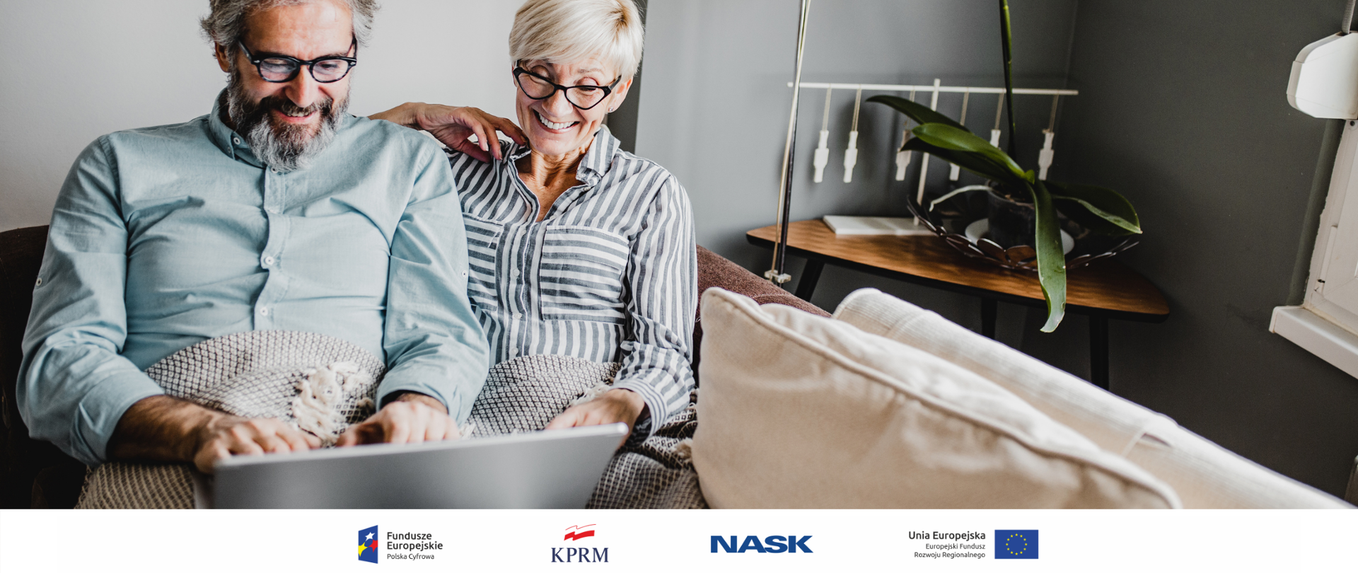 Dwoje dojrzałych, uśmiechniętych osób siedzi na kanapie i korzysta z laptopa.