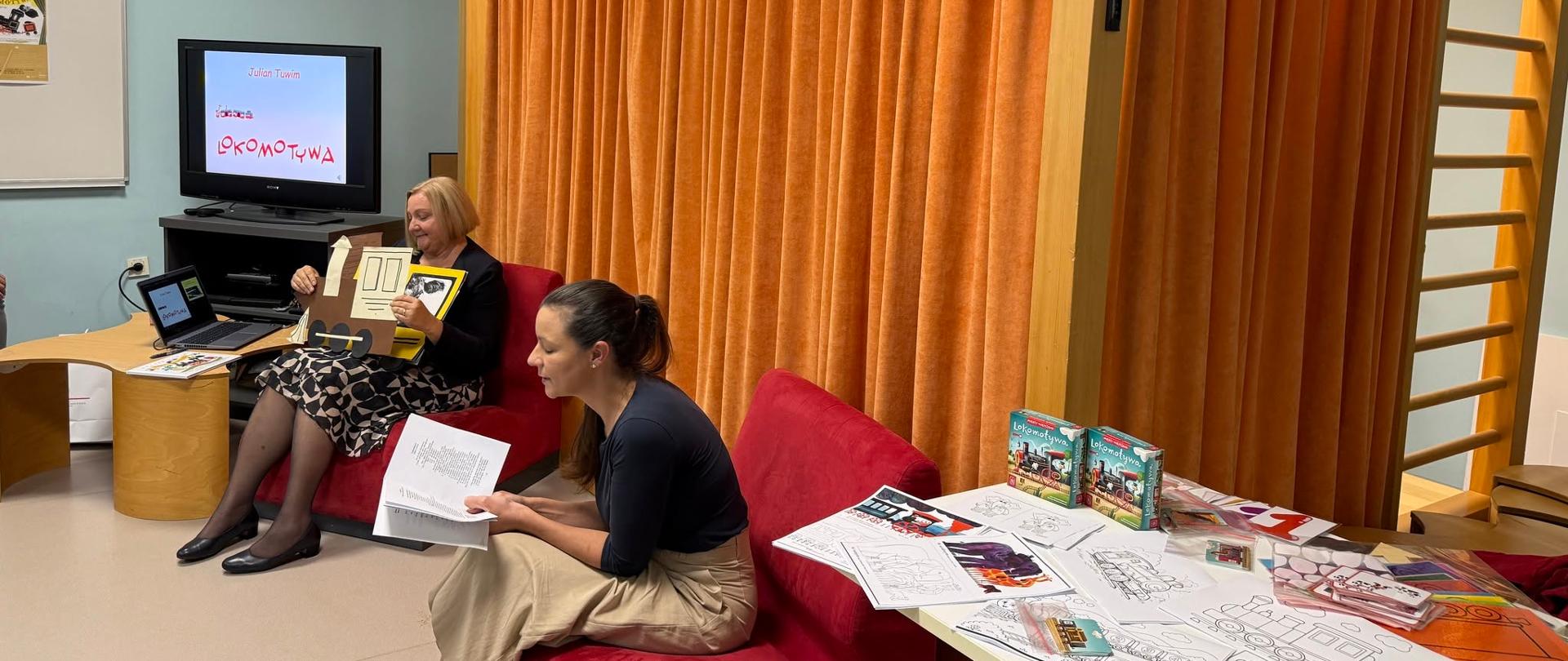 Izvajalki pripravljata gradivo za srečanje z otroki. Na zaslonu je naslov Lokomotywa Juliana Tuwima, na mizi pa so pobarvanke, ilustracije vlakov in drugo gradivo za ustvarjalne dejavnosti.