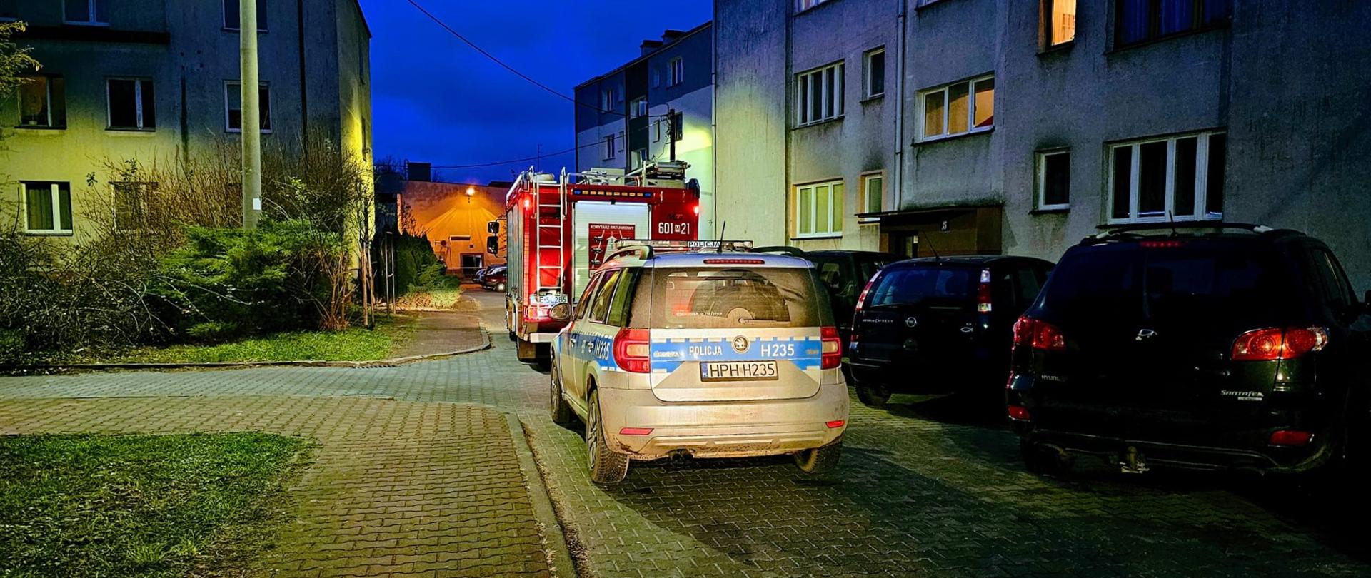 Nocna scena przedstawiająca ulicę osiedlową w Polsce. Na pierwszym planie widoczny jest zaparkowany radiowóz policyjny, a przed nim czerwony wóz strażacki. Po obu stronach ulicy znajdują się wielopiętrowe budynki mieszkalne z oświetlonymi oknami.