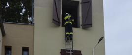 Szkolenie podstawowe strażaków-ratowników OSP. Plac komendy powiatowej w Rawiczu. czterech strażaków - kursantów wykonuje ćwiczenie z użyciem drabiny przystawionej do wspinalni. Jeden z ćwiczących stoi na szczycie drabiny - na wysokości drugiego piętra. Pomiędzy jego nogami znajduje się wąż, który wraz z prądownicą przewieszony jest przez prawe ramię strażaka. 