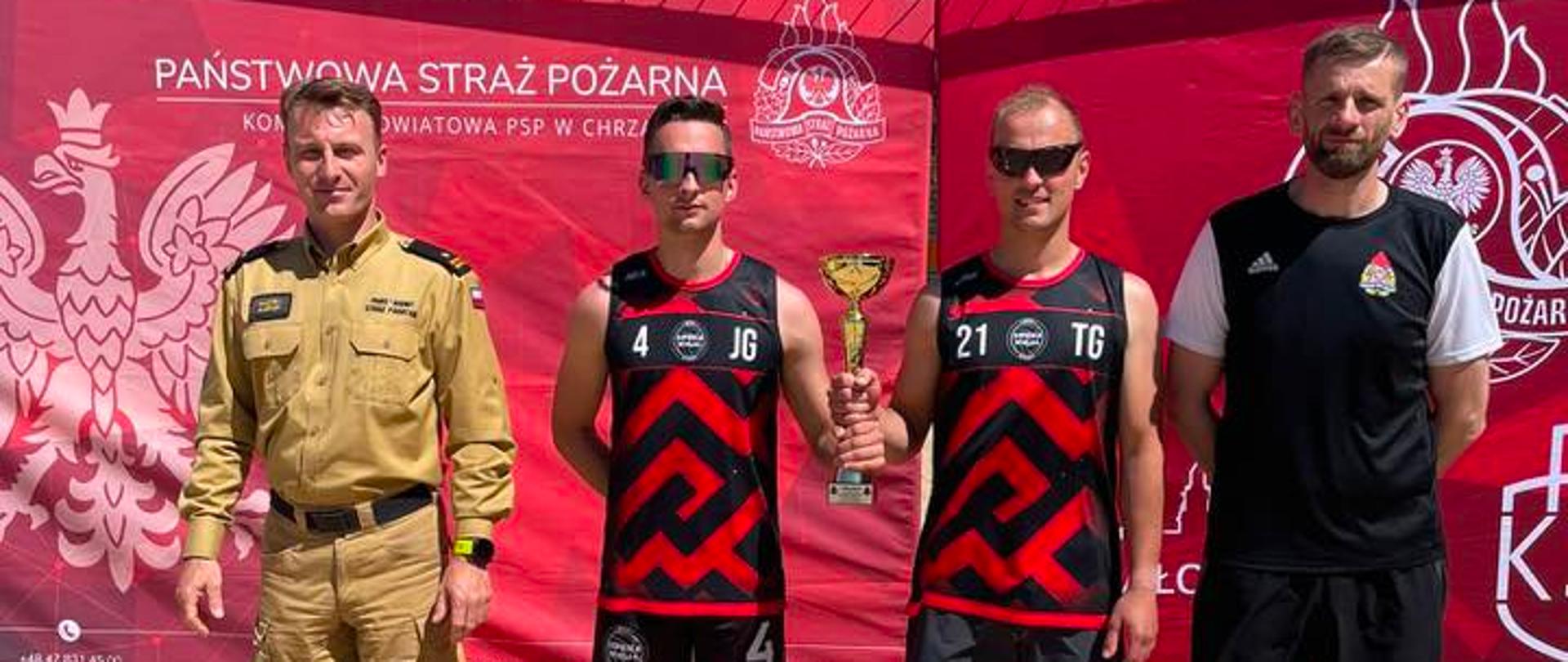Zdjęcie zwycięskiej drużyny z pucharem. Zwodnicy tworzący zwycięską drużynę trzymają puchar za zwycięstwo w zawodach. Mają na sobie czerwono-czarne stroje sportowe. Siatkarze stoją na piasku, bez obuwia. Mają założone ciemne okulary. Z lewej strony zawodników stoi strażak ubrany z piaskowe ubranie służbowe, z prawej natomiast osoba z grona organizatorów zawodów ubrana w czarny sportowy strój z logiem PSP na koszulce. Wszystkie te osoby stoją na tle czerwonego banneru, na którym widać z lewej strony orła, u góry napis Państwowa Straż Pożarna a po prawej stronie logo PSP. Elementy banneru są częściowo zasłonięte przez siatkarzy, strażaka oraz organizatora. 