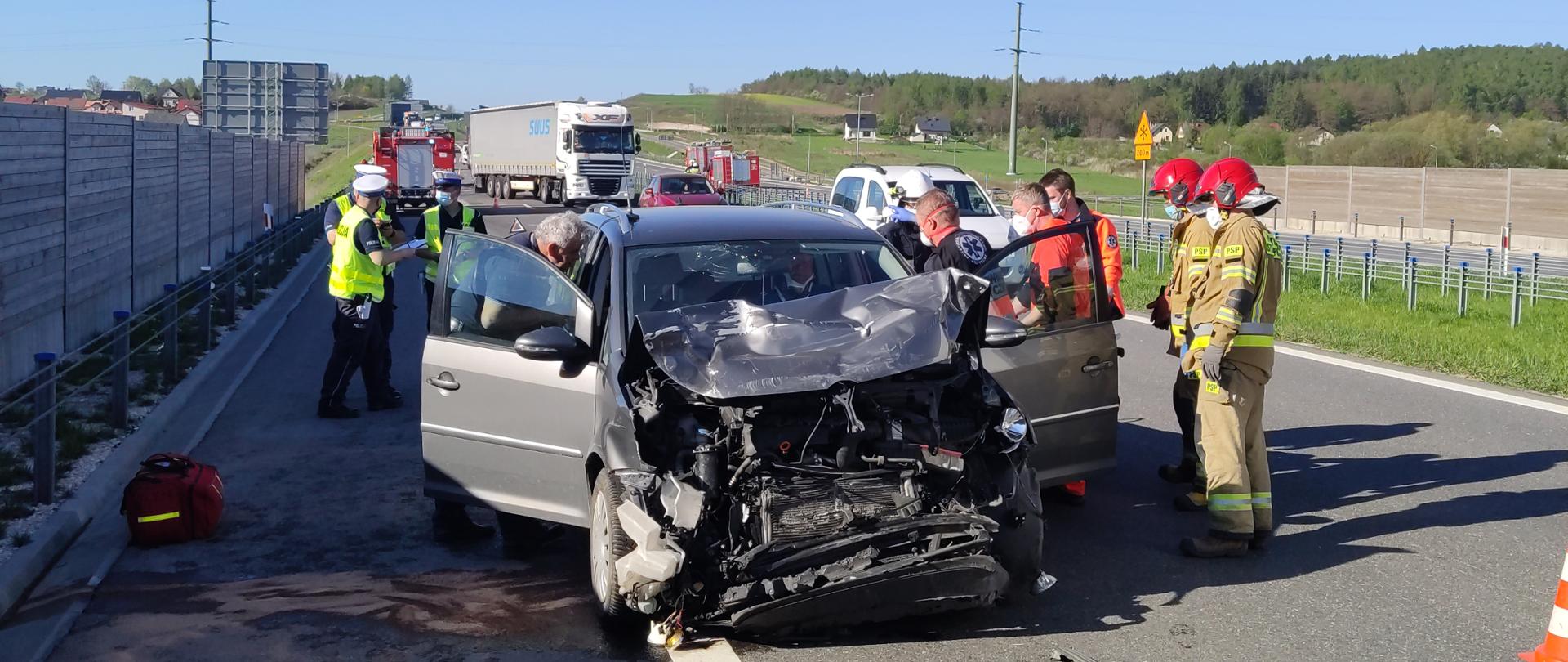 Zdjęcie przedstawia uszkodzony samochód marki Toyota Avensis stojący na poboczu drogi ekspresowej. Obok samochodu znajdują się strażacy. Droga jest zablokowana.