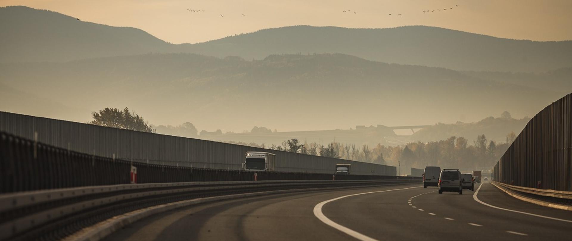 Fotografia dwujezdniowej drogi ekspresowa S1. W tle widoczne góry. W górnej części fotografii widoczne niebo z kluczem przelatujących ptaków. Po jezdniach w obu kierunkach poruszają się pojazdy. 