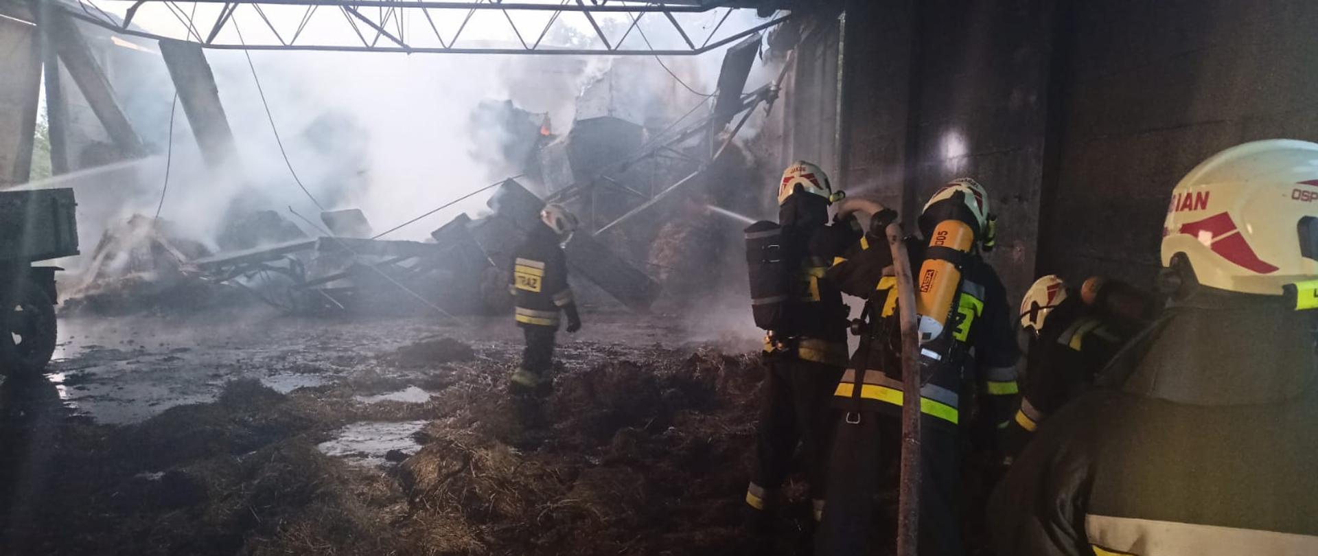 Wnętrze stodoły po pożarze. W tle fragment stodoły nad którym zawalił się dach. Na pierwszym lanie po prawej widać kilku strażaków OSP, dwaj z nich trzymając wąż podaje prąd wody w kierunku gruzowiska. Strażacy w hełmach koloru białego, i granatowych ubraniach specjalnych. 