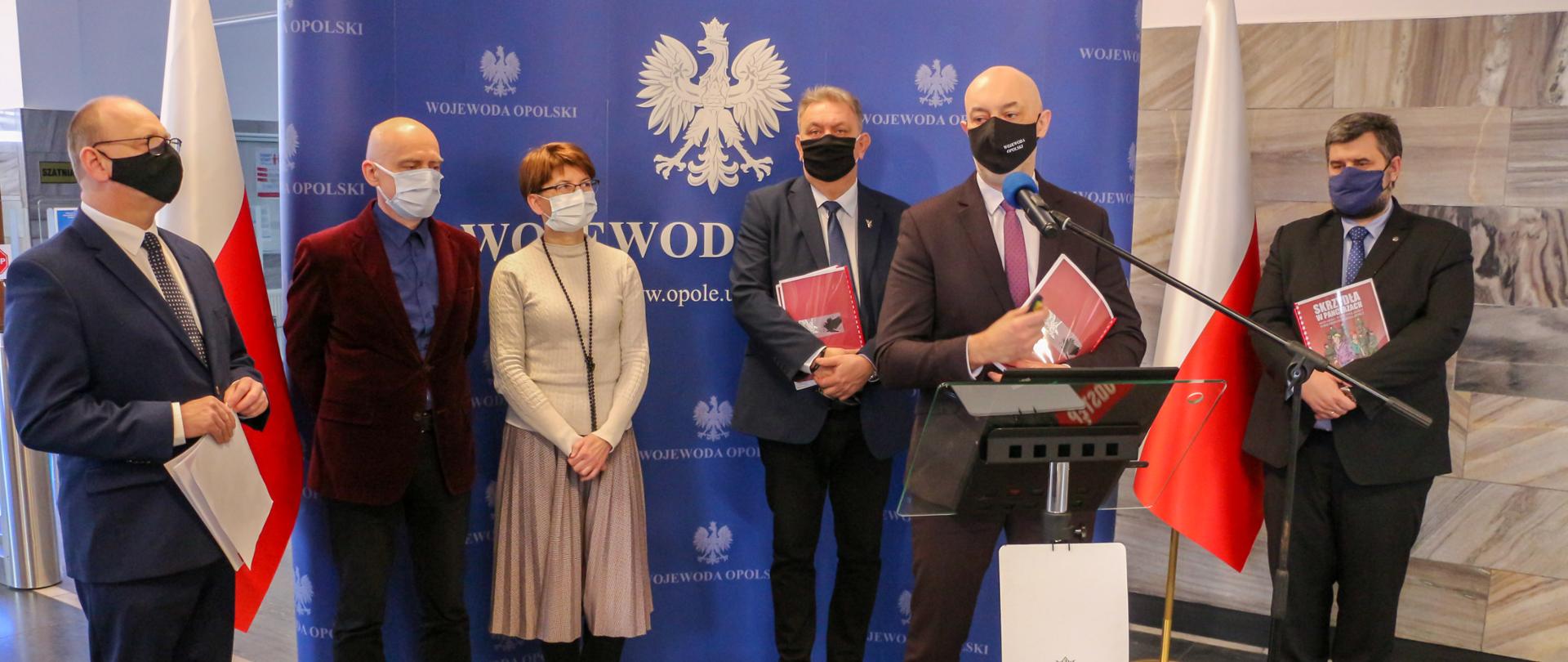 Konferencja prasowa wojewody opolskiego . Na zdjęciu na pierwszym planie wojewoda Adrian Czubak, za nim stojący na tle ścianki granatowej z napisem "Wojewoda Opolski", partnerzy Niepodległej. Od lewej dyrektor Biura Wojewody Opolskiego, ubrany w granatowy garnitur; dyrektor EkoStudio - ubrany w bordową marynarkę i ciemne spodnie ; dyrektor CMJW w jasnej kreacji; dyrektor brzeskiego muzeum - w garniturze, trzymający w dłoni dokument. Za wojewodą po prawej stornie stoi również ubrany na czarno dyrektor Instytutu Śląskiego, który spogląda w dal, a w dłoniach trzyma komiks. Po bokach kadru biało-czerwone flagi. 