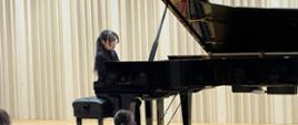 Na zdjęciu czarny fortepian, a przy nim kobieta brunetka ubrana na czarno. Tło obrazka w odcieniach beżu. Na dole zdjęcia fotele, a na nich siedzi zgromadzona publiczność.