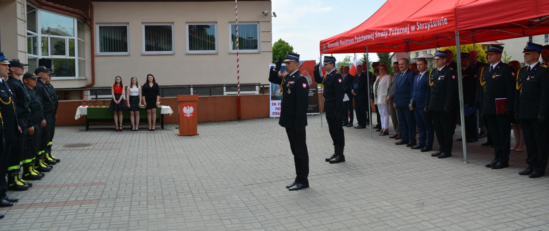 Zdjęcie wykonane na zewnątrz. Po lewej stronie zdjęcia dwóch oficerów Państwowej Straży Pożarnej salutując pododdziałowi. W tle stoją goście uczestniczący w uroczystości.