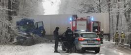 Po lewej widać samochód ciężarowy, znajduje się przy drzewie, na środku radiowóz Policji, za nim naczepa pojazdu i samochód strażacki