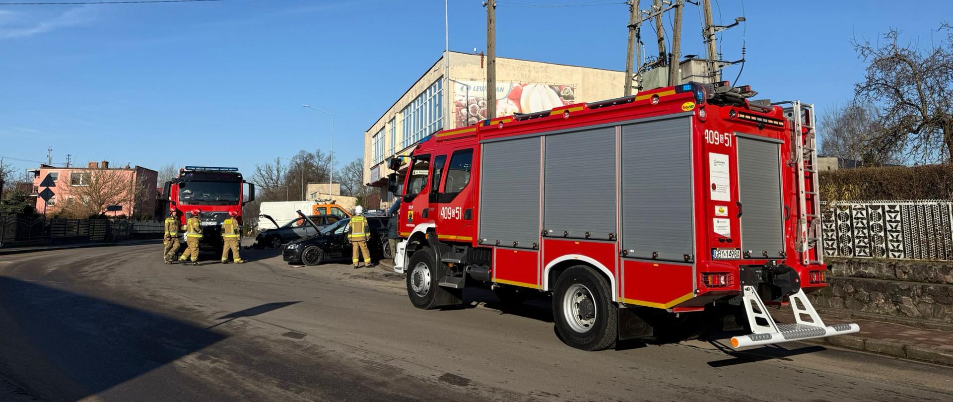 Czerwony wóz strażacki i strażacy w ubraniach specjalnych koloru piaskowego podczas działań.