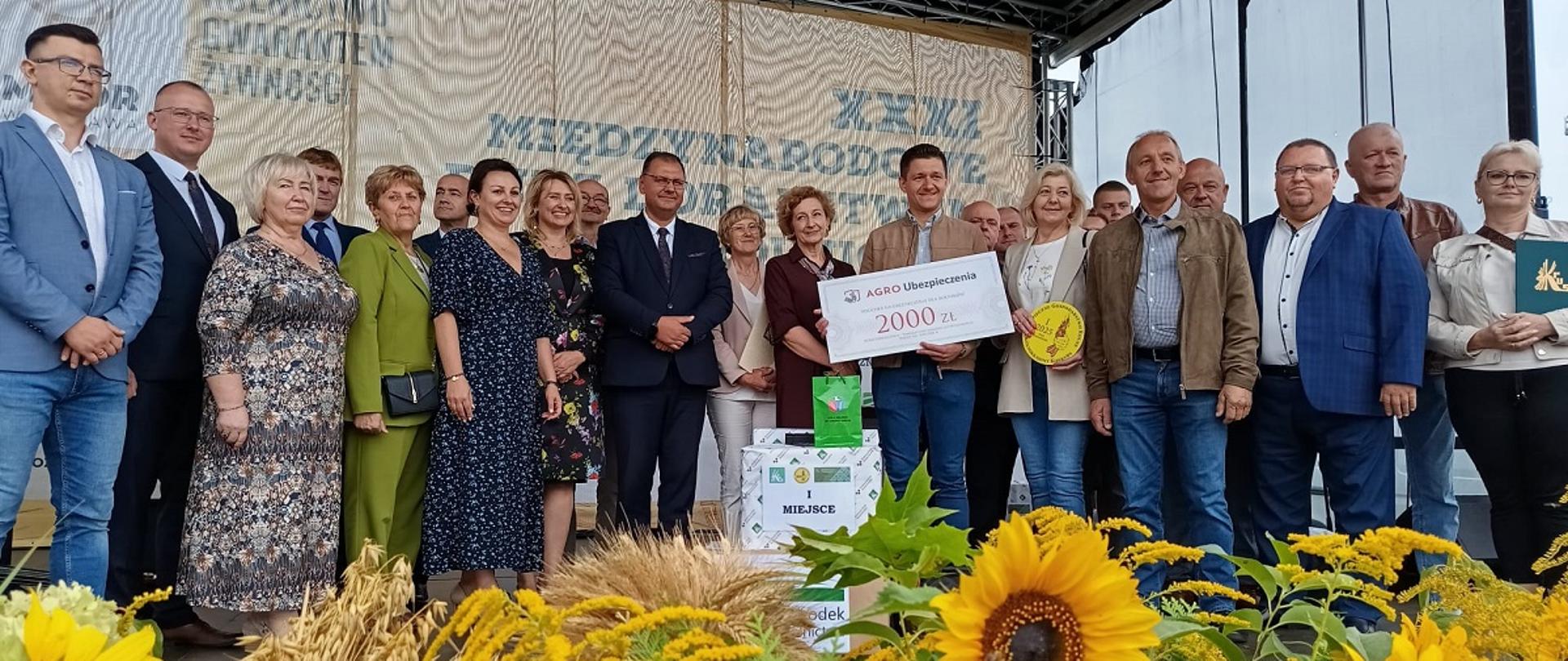 Grupa dwudziestu trzech osób stojących na scenie pozuje do zdjęcia podczas wydarzenia plenerowego. Za nimi w tle baner z napisem: „Międzynarodowe Dni z Doradztwem Rolniczym”. Mężczyzna stojący na środku trzyma w rękach duży symboliczny czek z napisem AGRO Ubezpieczenia” i kwotą 2000 zł. Przed nim stoją trzy kartony owinięte w papier, z przodu najbardziej widocznego kartonu przyklejona jest biała kartka z napisem I MIEJSCE, na kartonie stoi zielona torebka prezentowa. Kobieta stojąca po jego prawej stronie trzyma żółty okrągły znaczek z logo BGR. Kobieta stojąca pierwsza z prawej trzyma przed sobą teczkę z logo KRUS. Z przodu scena udekorowana jest żółtymi kwiatami słonecznika, nawłoci i kłosami zbóż.