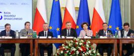 Przy stole siedzi grupa elegancko ubranych osób, wśród nich prezydent Duda, przed nimi wielki bukiet biało-czerwonych kwiatów, za nimi flagi Polski i UE. Z boku tablica z napisem Rada Dialogu Społecznego.