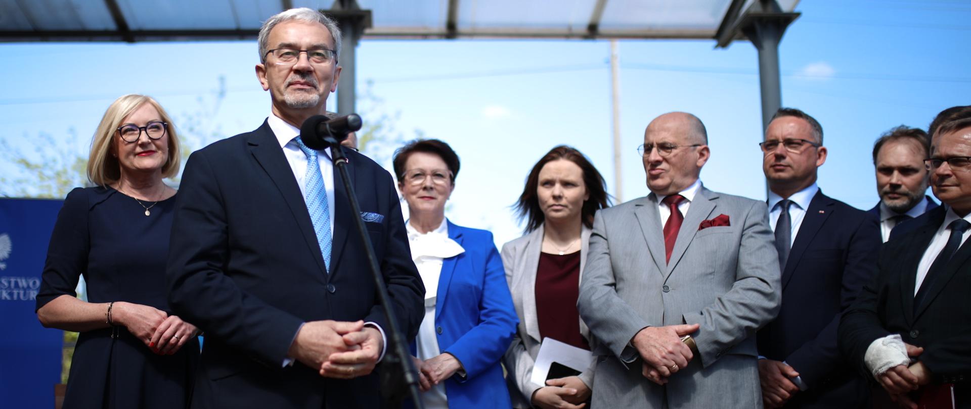 Minister Jerzy Kwieciński stoi przy mikrofonie. Za nim stoją pozostali uczestnicy przemówienia. 