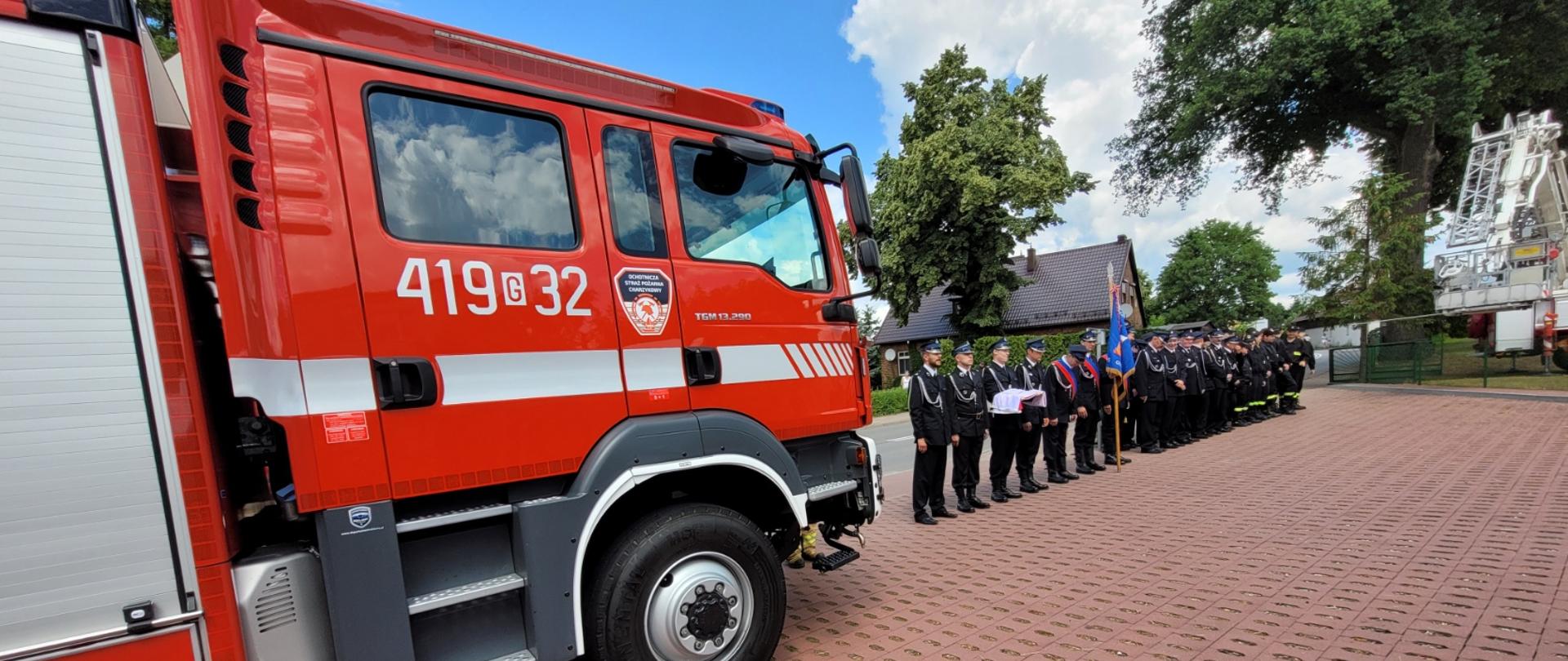Strażacy OSP Charzykowy stoją z flagą, ze sztandarem obok nowego samochodu gaśniczego.