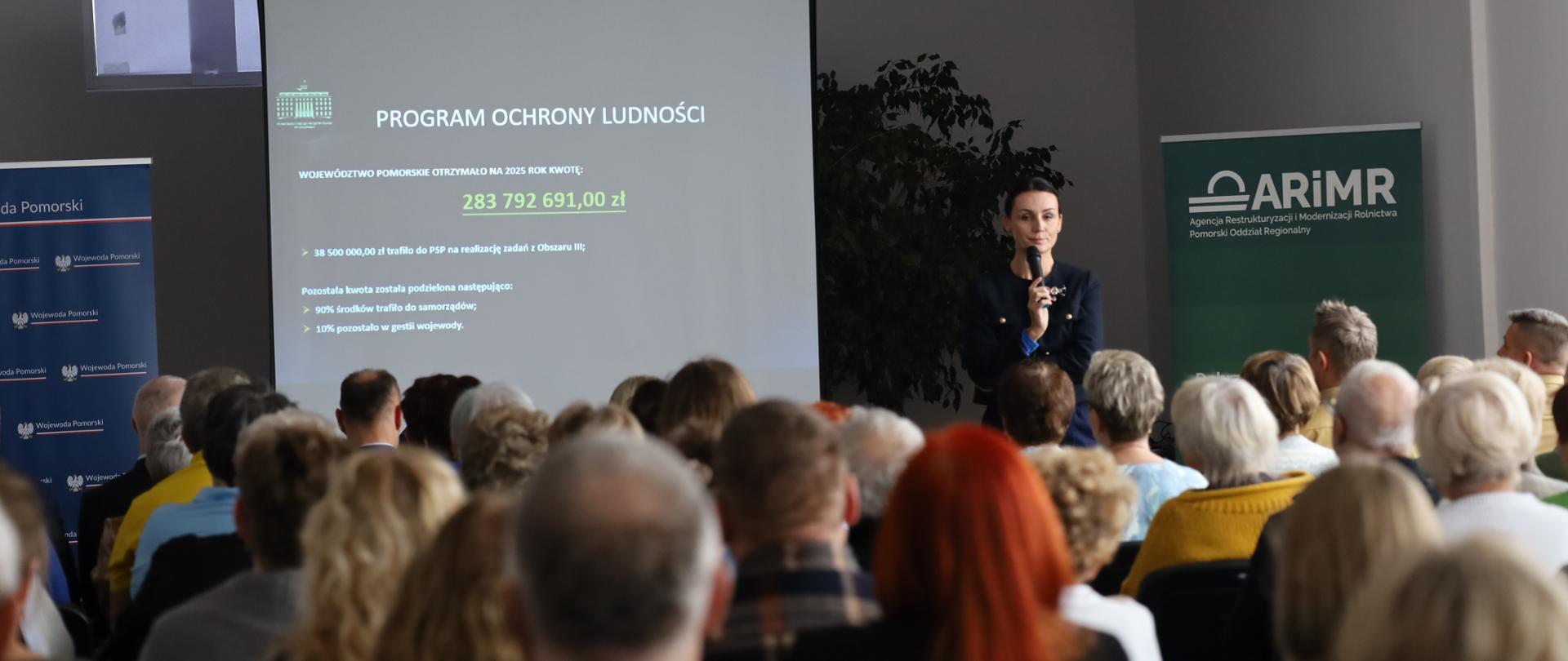 Na zdjęciu widać grupę ludzi (słuchaczy szkolenia). W tle widać kobietę (wojewodę). Kobieta prowadzi prezentację na temat programu ochrony ludności o obrony cywilnej.