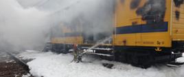 Strażak przyklękając w pianie gaśniczej podaje prąd wody do wagonu kolejowego dogaszając pożar i chłodząc butle.