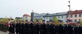 Strażacy stoją w dwuszeregu na placu wyłożonym kostką. Za nimi widać wysokie budynki. Obok strażaków z lewej strony stoją żołnierze.