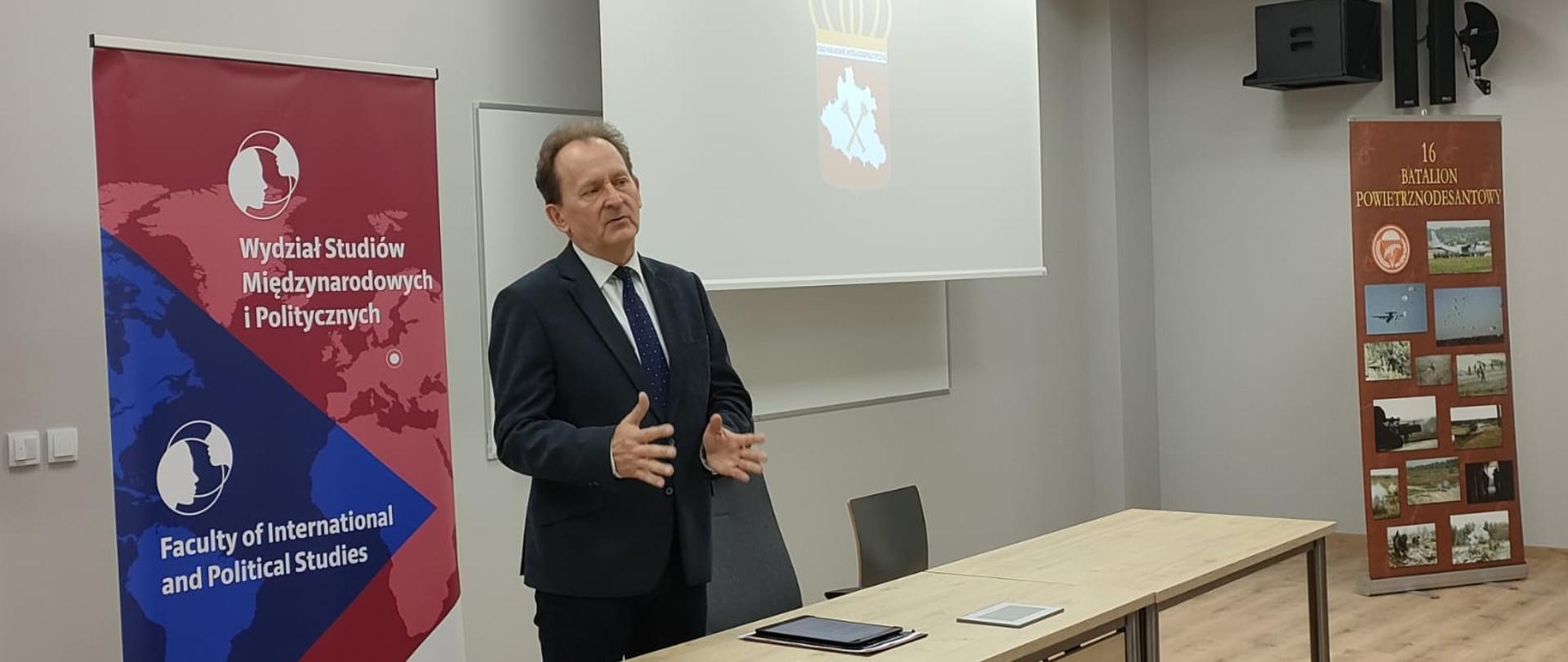 Minister Bernacki stoi za stołem i mówi do słuchaczy.