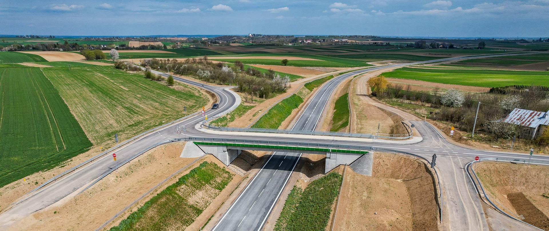 Łącznik północny DK74 - na zdjęciu z drona jednojezdniowa droga asfaltowa, na około zielone pola. Nad drogą wiadukt, do którego doprowadzone są drogi lokalne 